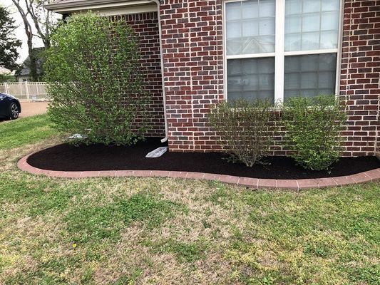 Mulched garden bed with brick border and bushes in front of a brick house, with green grass.