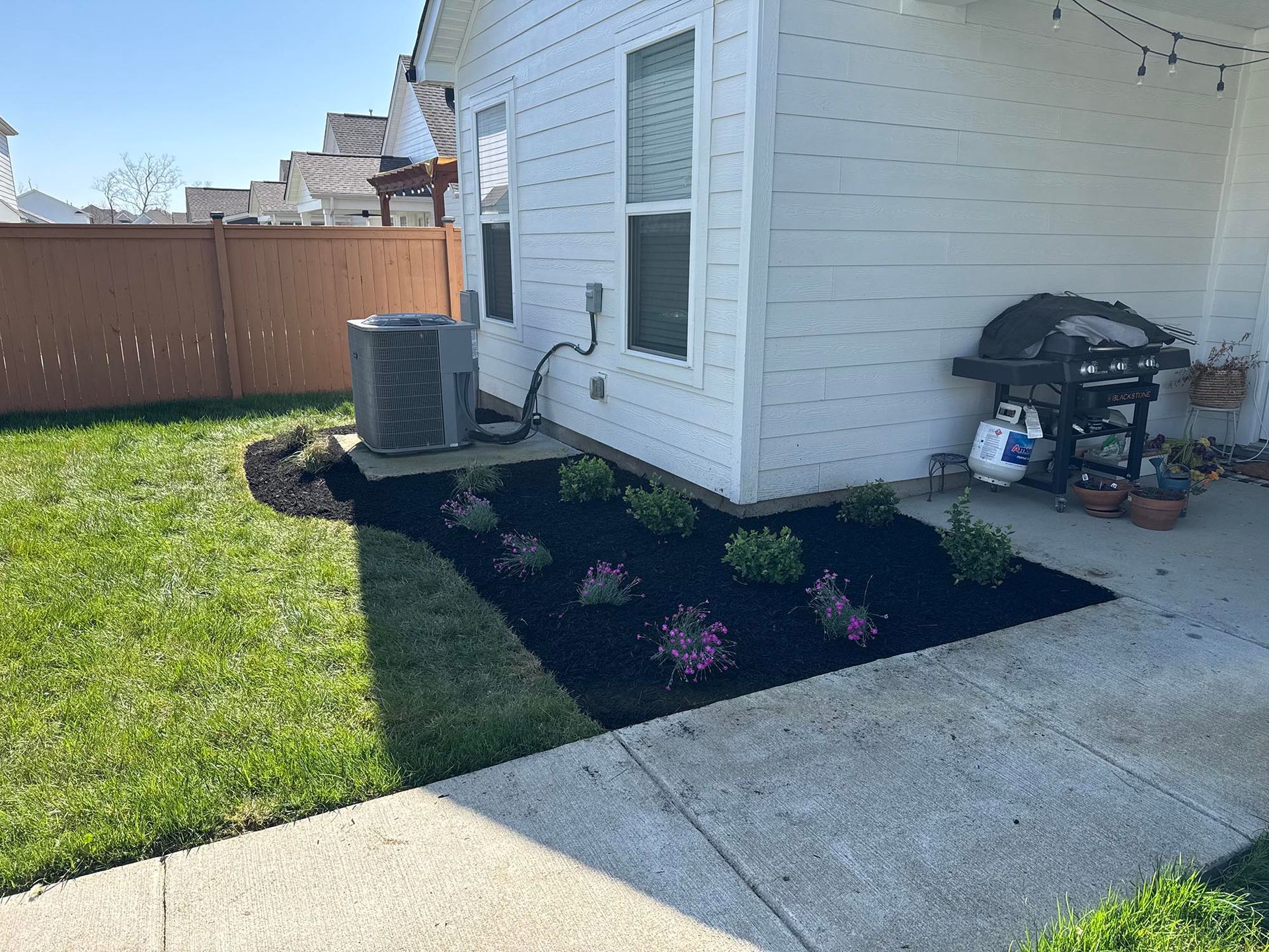 Backyard with flower bed, AC unit, grill, and white house.  Green grass, brown fence, and black mulch are visible.