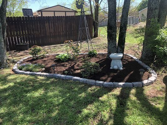 Landscaped garden bed with a stone border, mulch, plants, and a white statue. Trees and a wooden fence in the background.