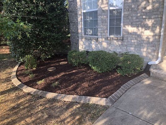 A landscaped flower bed with dark mulch, bordered by brick, next to a brick house and sidewalk.