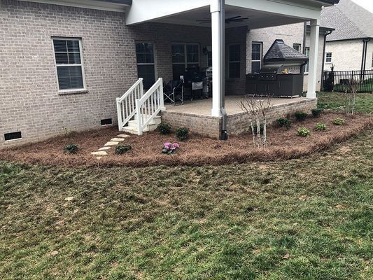 Backyard with a brick house, patio, and a flower bed edged with pine straw.