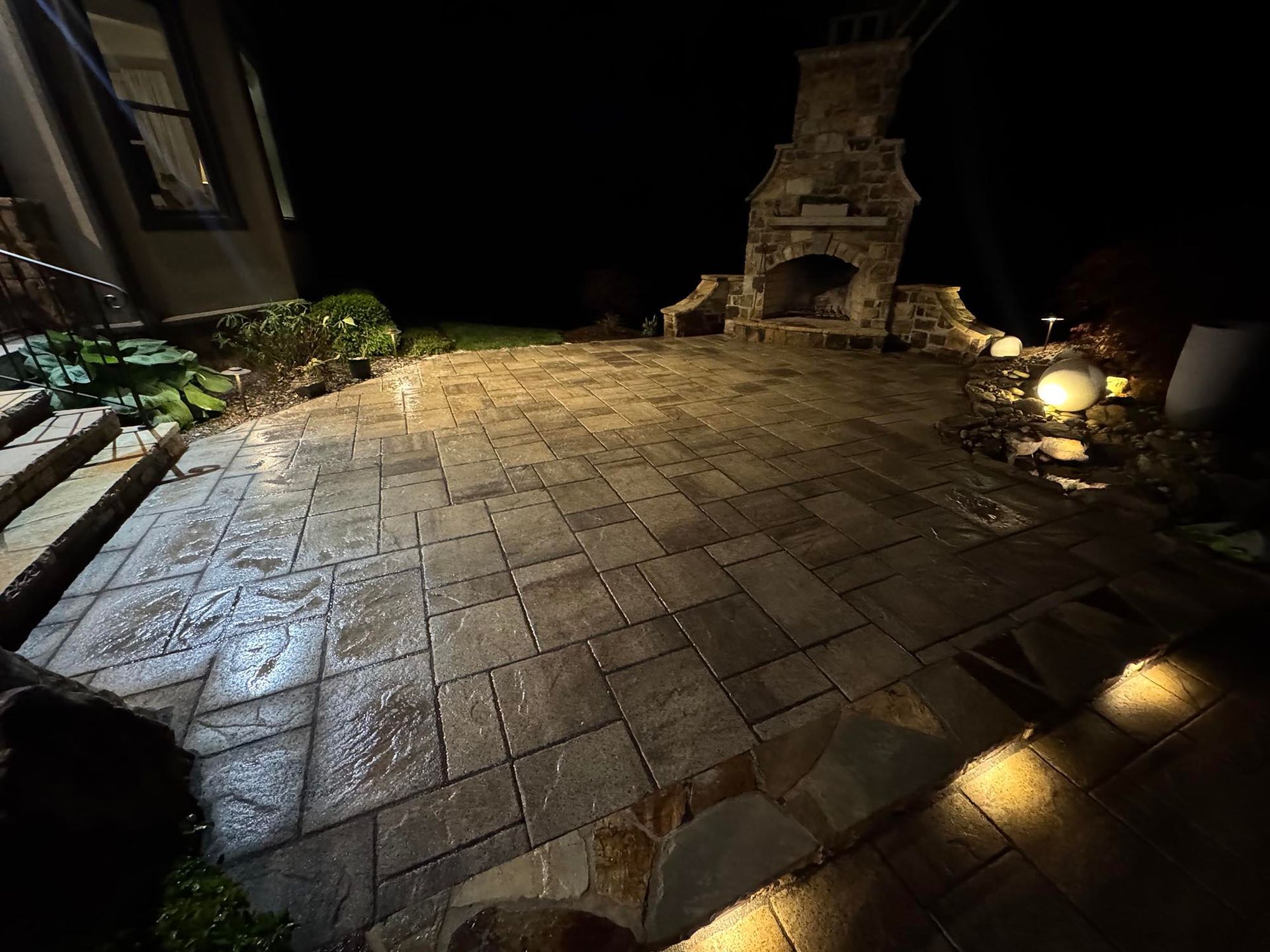 Nighttime view of a paved patio with an outdoor fireplace, illuminated by ground lights and house lights.