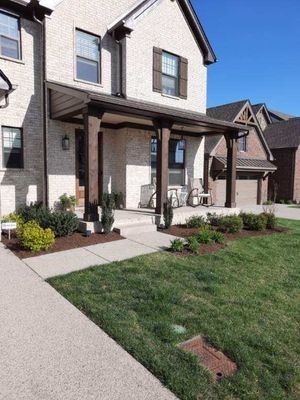 Beige brick house with a dark brown porch, front yard with greenery, and blue sky.