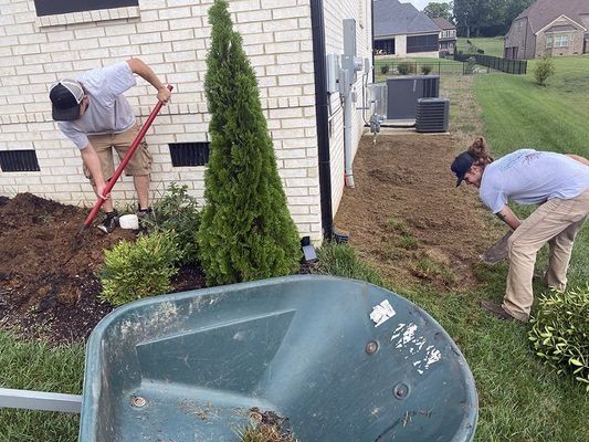 Two workers landscaping near a brick house, one using a shovel, the other working on the ground, wheelbarrow in foreground.