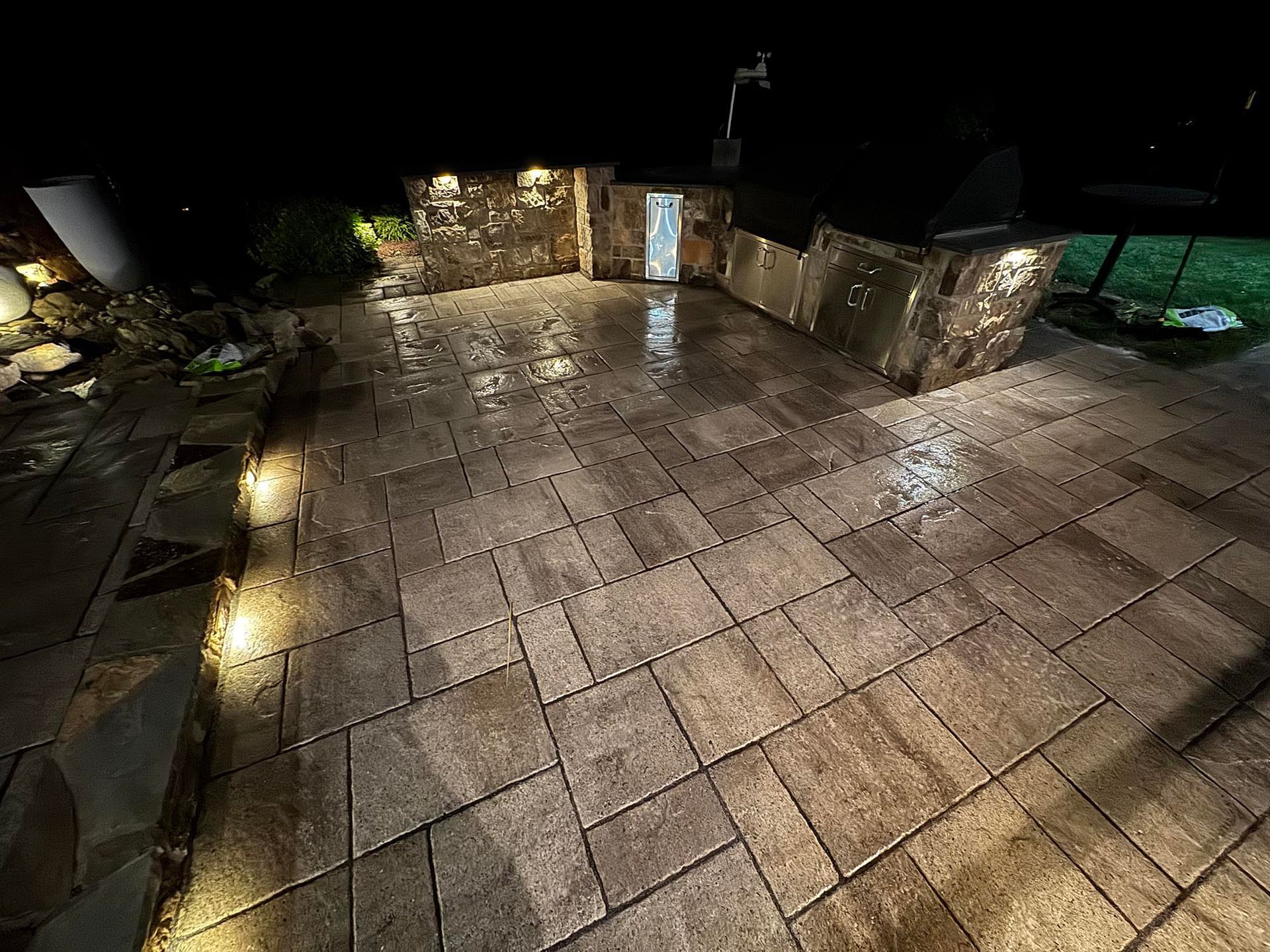 Stone patio at night, wet from rain, lit by spotlights. Built-in grill area with stone accents.