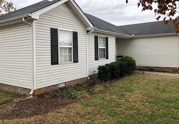 White house with black shutters, brown roof, and green bushes in front.
