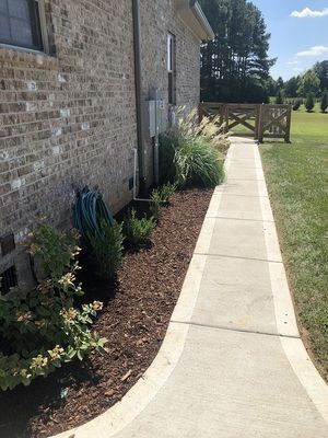 Sidewalk beside a brick building, with mulch-covered landscaping and a fence leading to a grassy field.