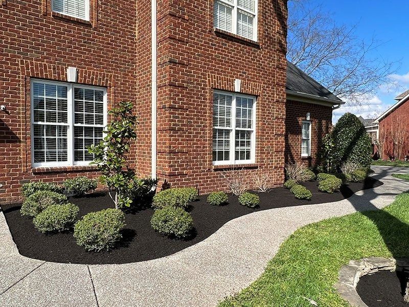 Brick house with white-framed windows, dark mulch beds, green shrubs, and a curved walkway.