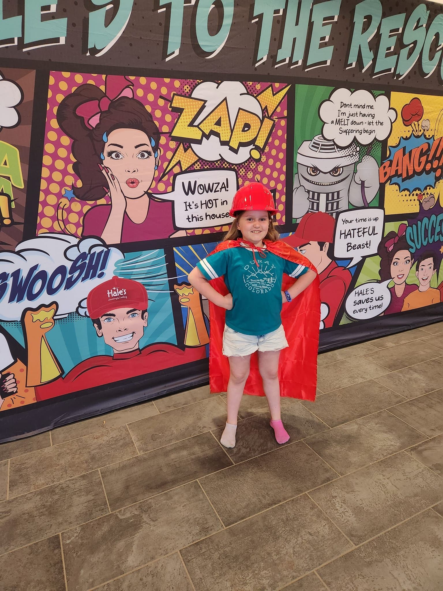A smiling child wears a red cape and hard hat, standing heroically in front of a colorful, comic book-style wall mural.