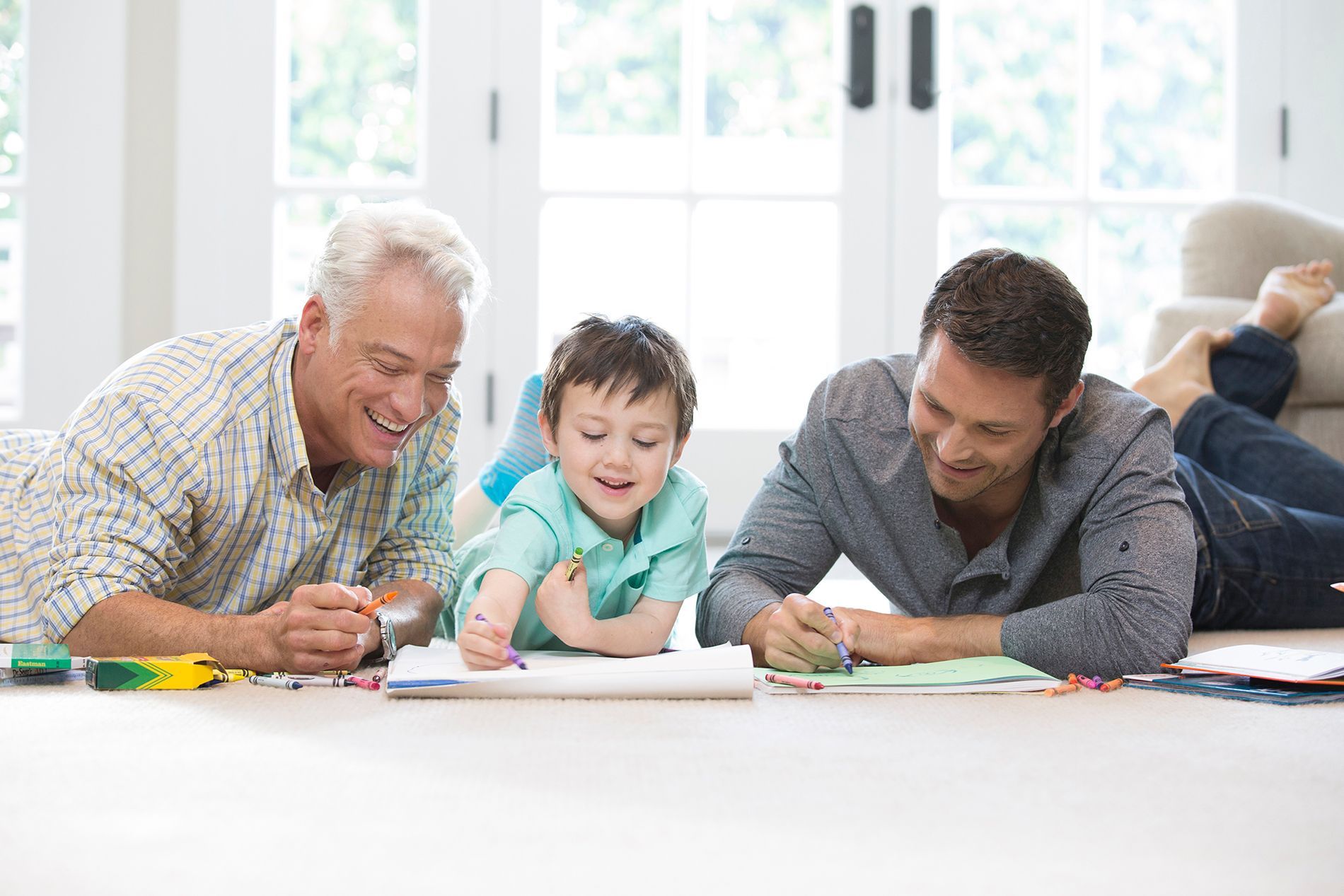 Two adults and a child lie on the floor together, happily coloring in notebooks in a bright, sunlit room.