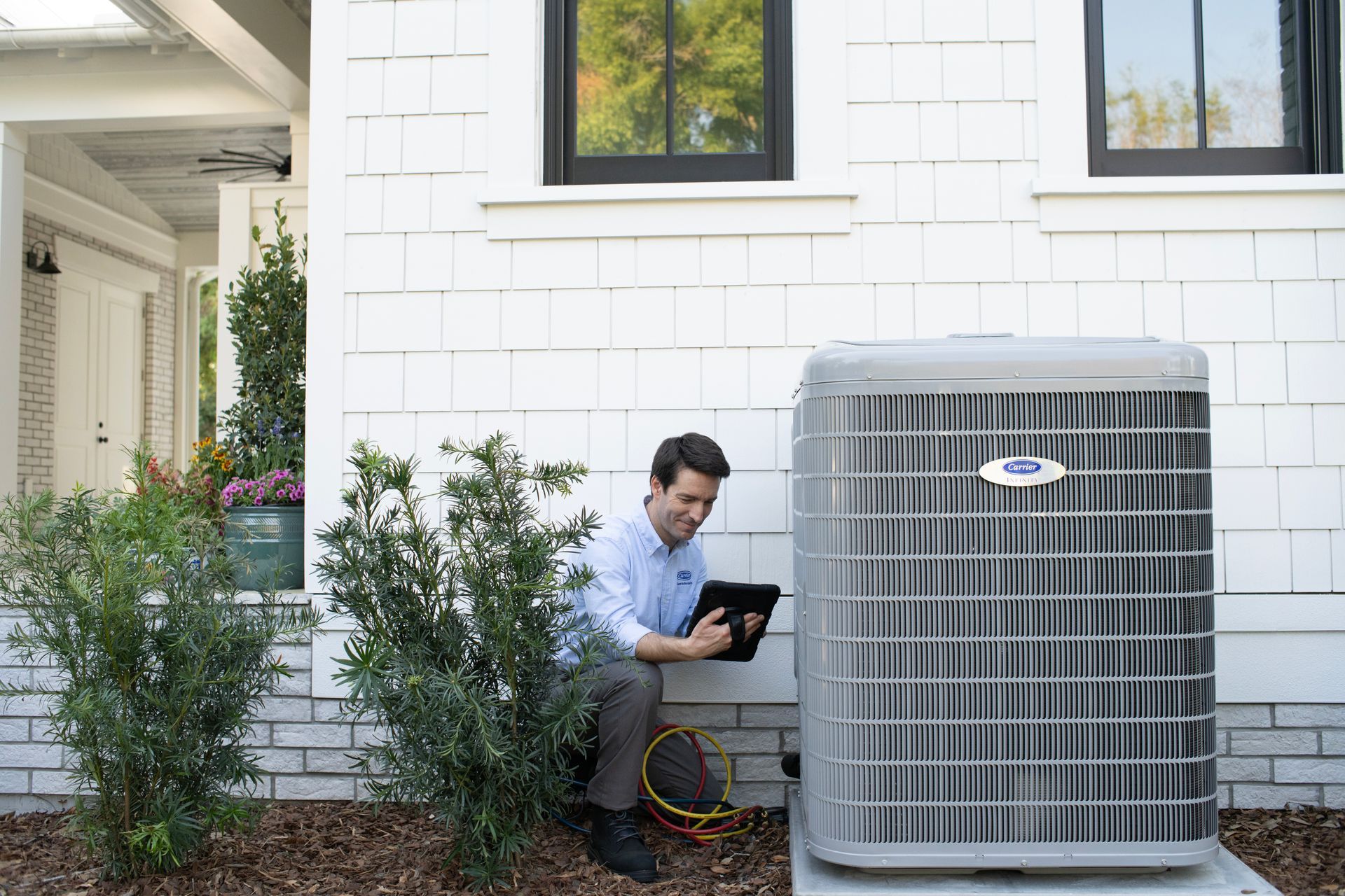 A technician kneels outside a house, using a tablet to inspect a gray air conditioning unit.