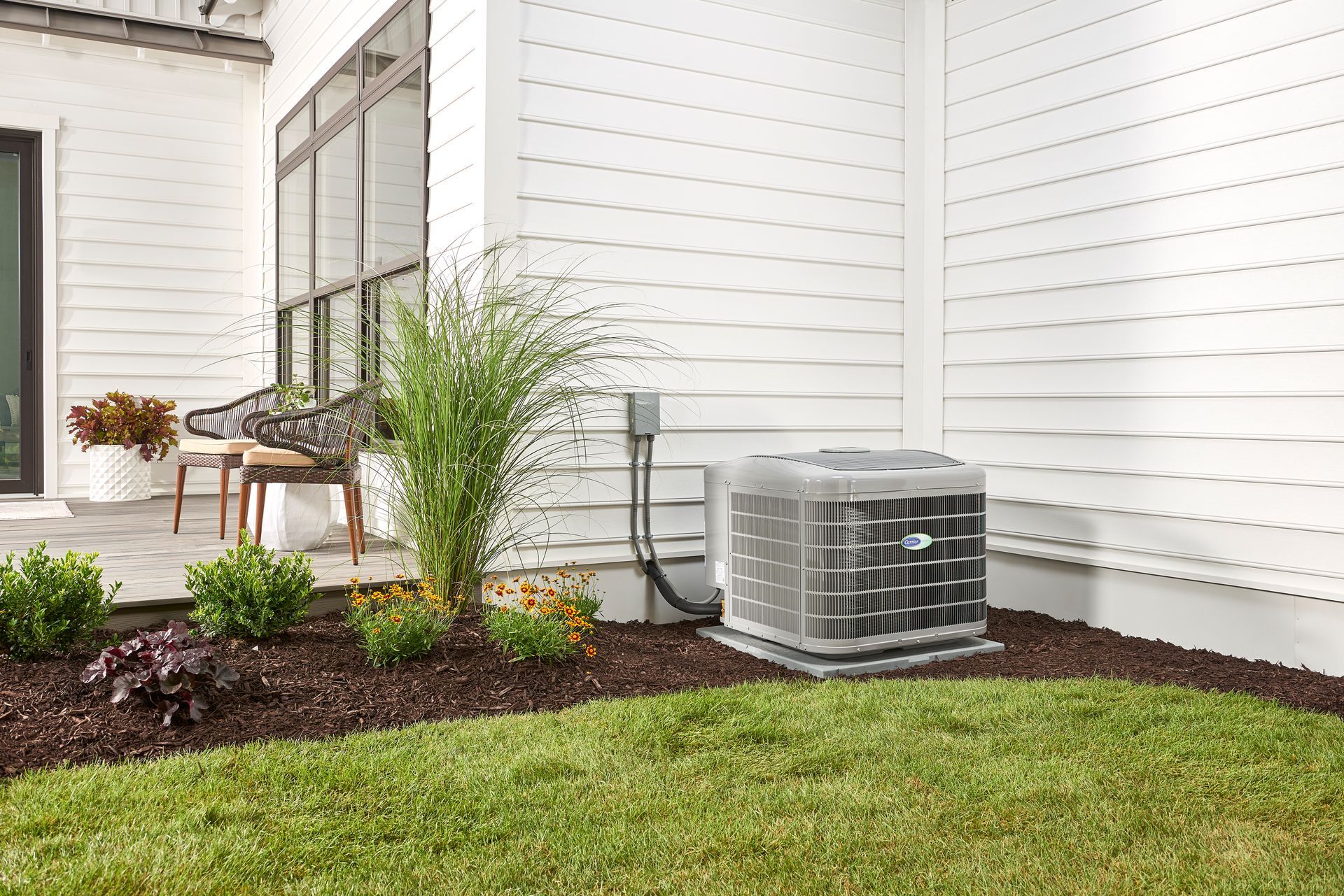 An outdoor HVAC unit sitting in a mulched flower bed next to a white house with a patio area.