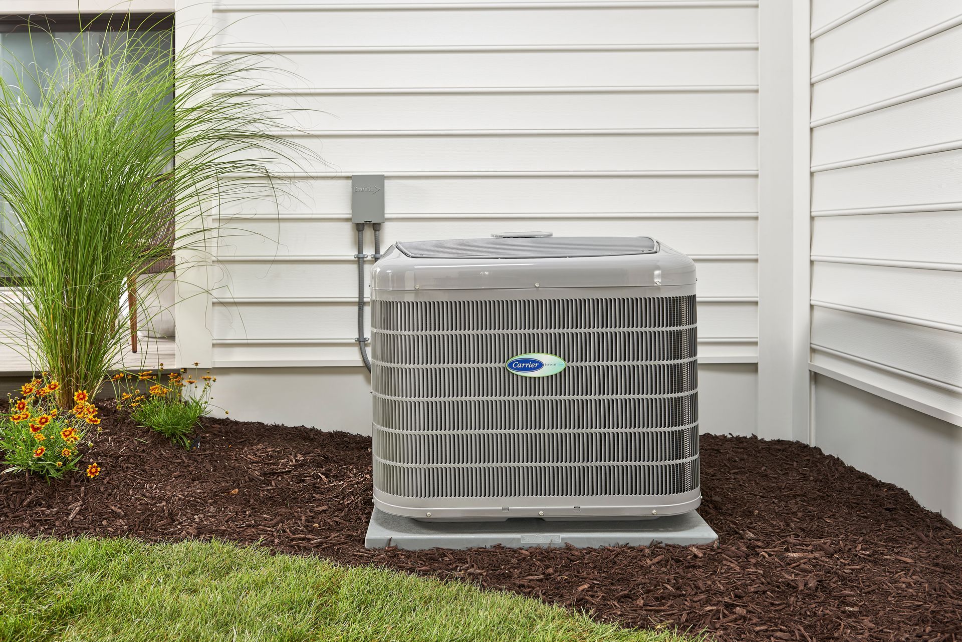 A gray residential air conditioning unit sitting on a concrete pad next to a white house with mulch and landscaping.