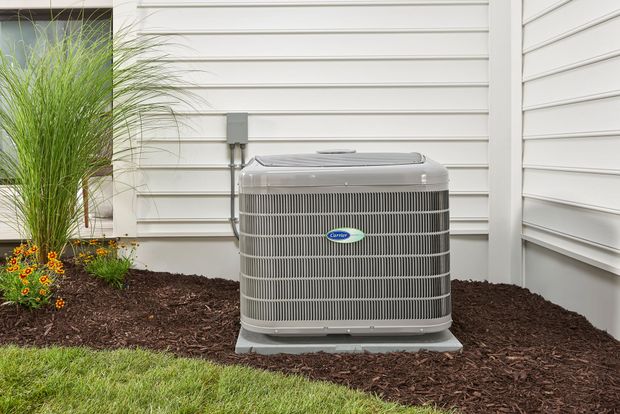 A gray residential air conditioning unit sitting on a concrete pad next to a white house with mulch and landscaping.