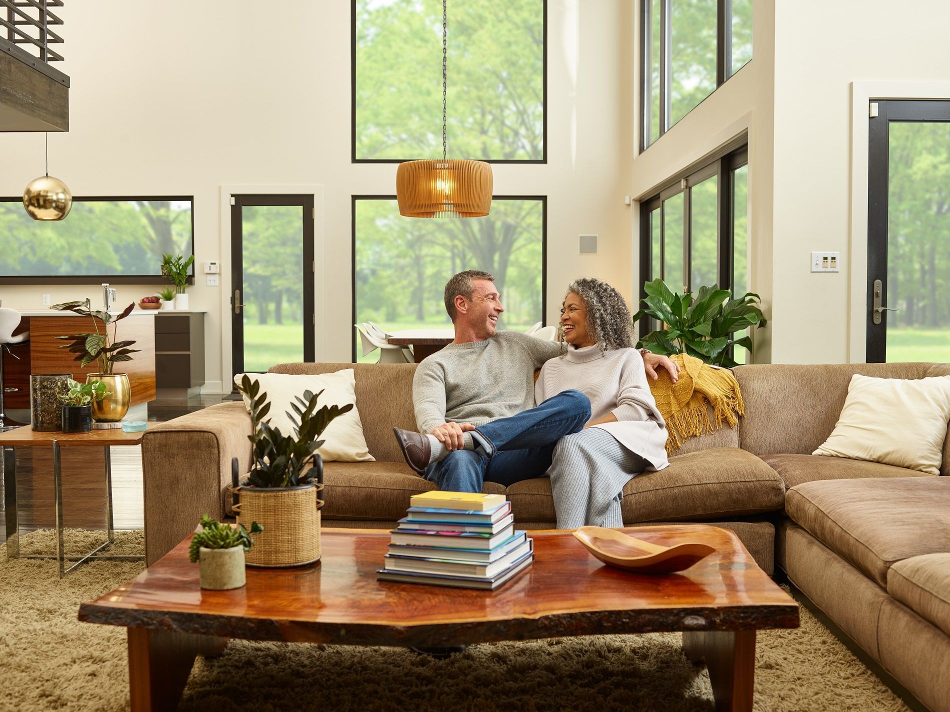 A couple sits together on a brown sofa in a sunlit living room with large windows, looking at each other and smiling.