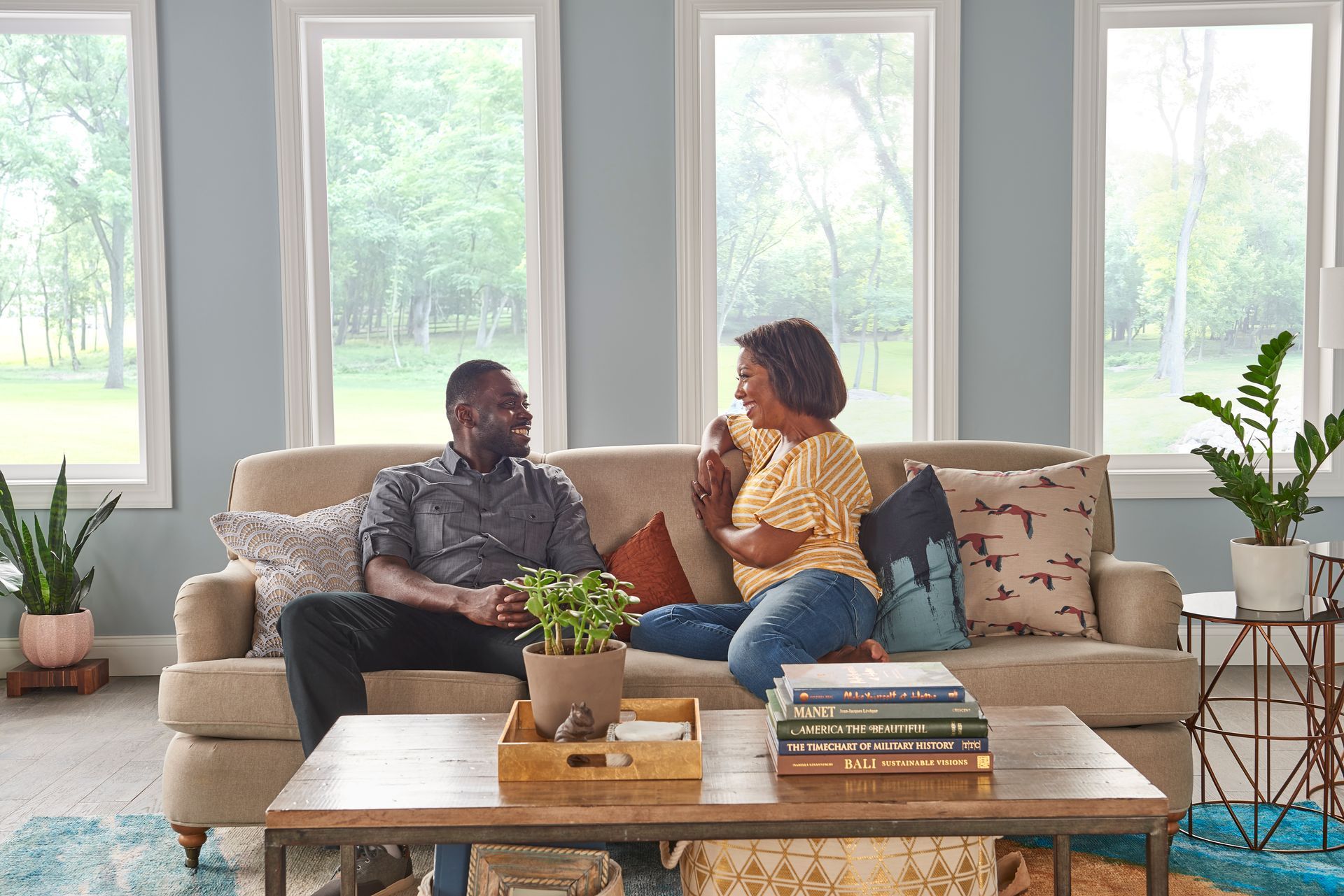 A couple sits on a couch in a sunlit living room, chatting and smiling at each other.