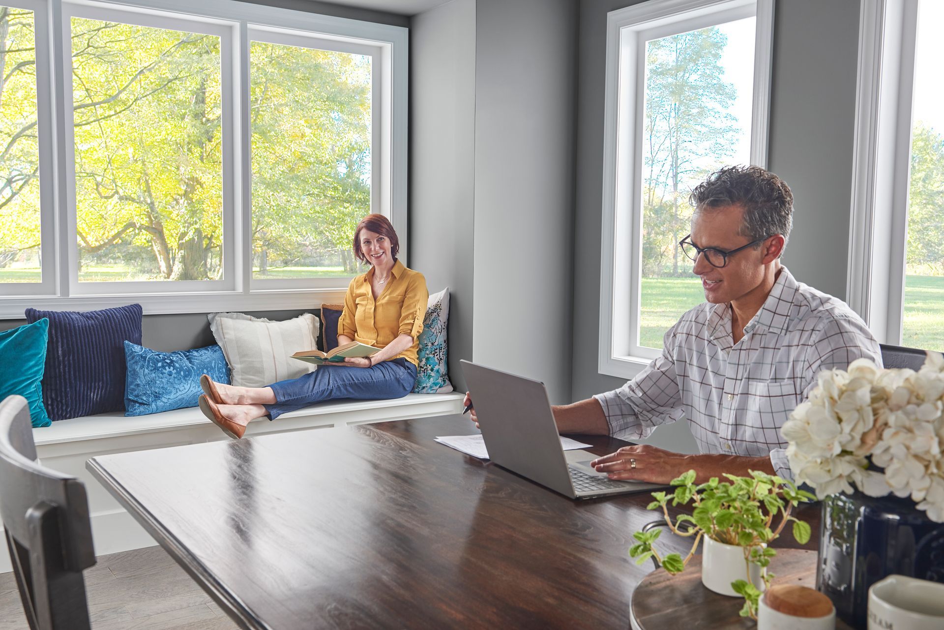 A person works on a laptop at a table while another sits on a nearby window seat in a bright, sunlit room.