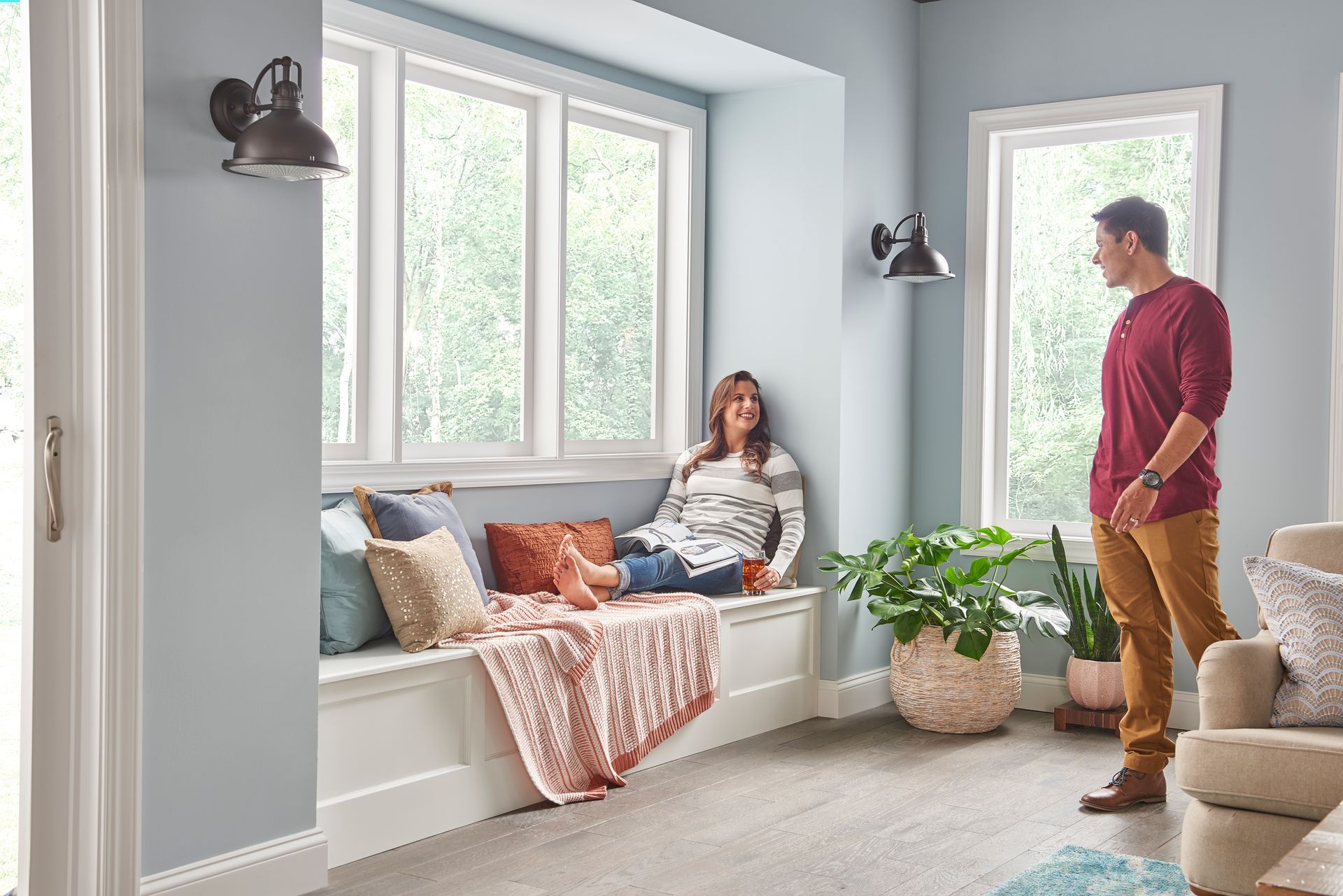 A person sits on a window seat filled with cushions and blankets, while another stands nearby in a sunlit, blue-walled room.