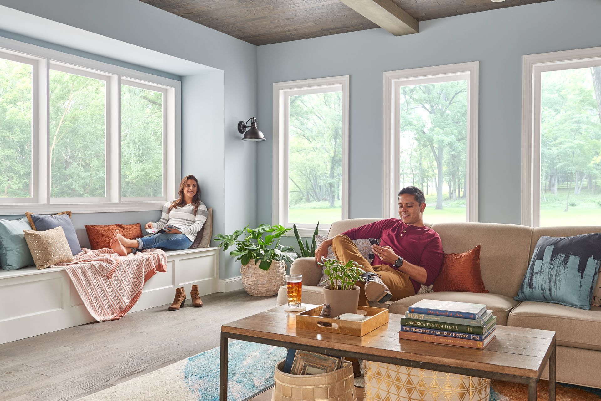 A room with large windows and light blue walls, featuring a person on a window seat and another relaxing on a beige sofa.