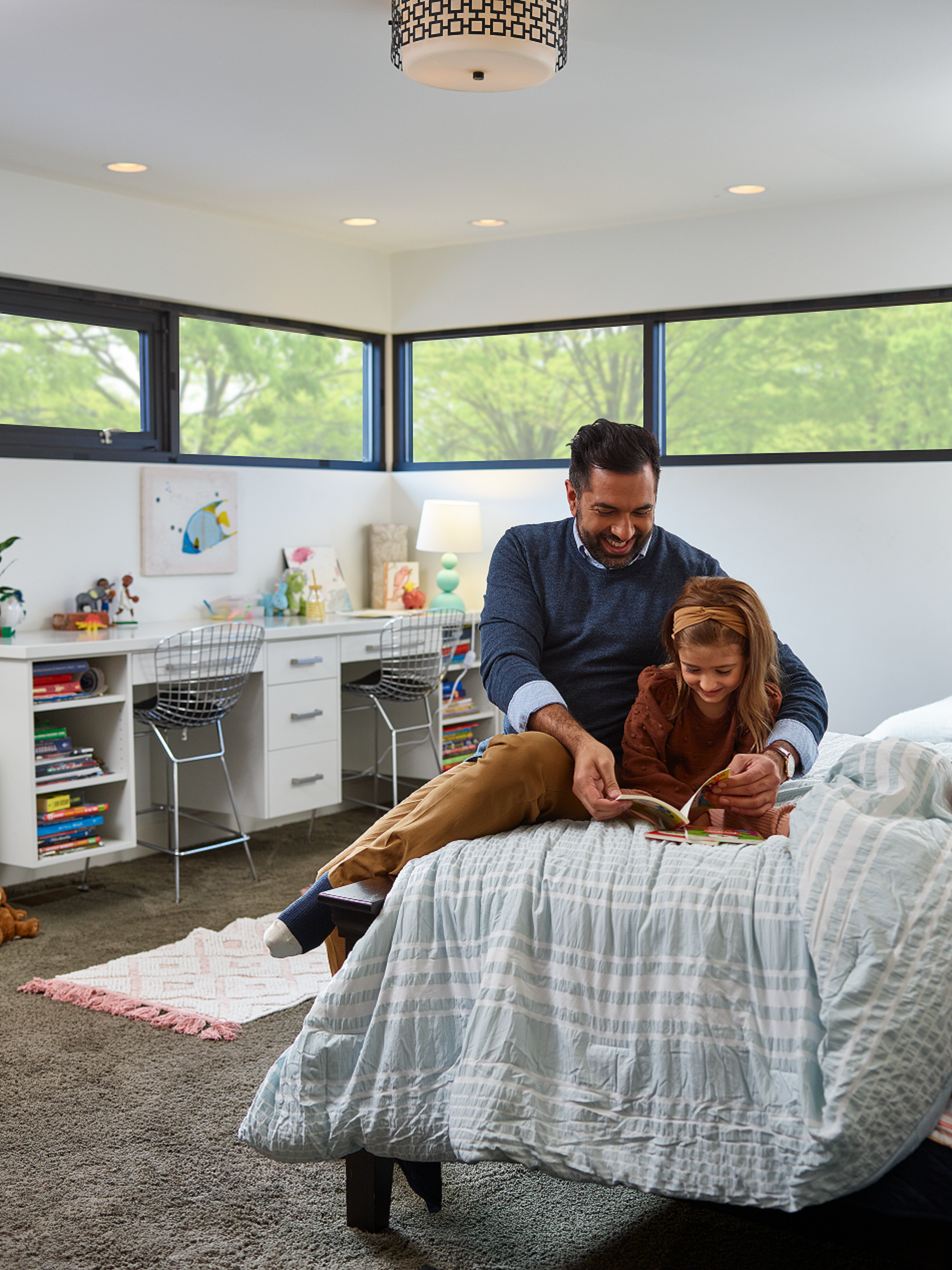An adult and child sit on a bed in a bright bedroom, looking at a book together in front of a white desk and large windows.