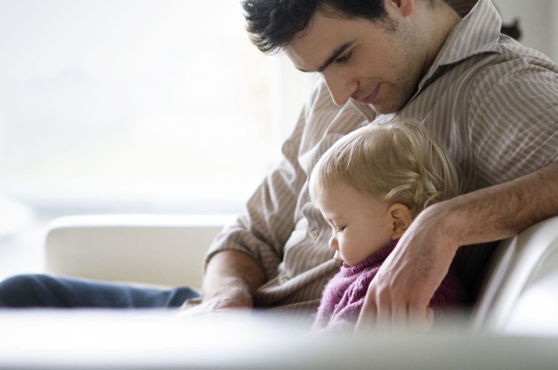 A parent in a striped shirt sitting on a white sofa, cradling a sleeping toddler wearing a purple sweater.
