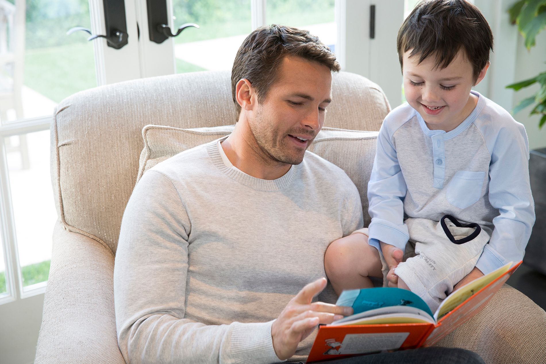 A person sits in an armchair reading a colorful book to a smiling child seated on their lap in a bright room.
