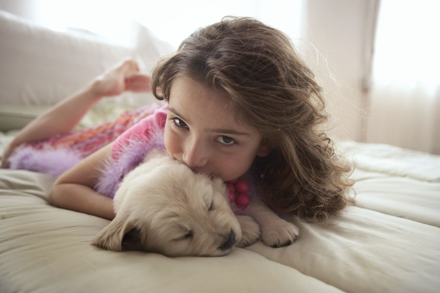 A person with wavy hair lying on a bed and resting their chin on a sleeping light-colored puppy.
