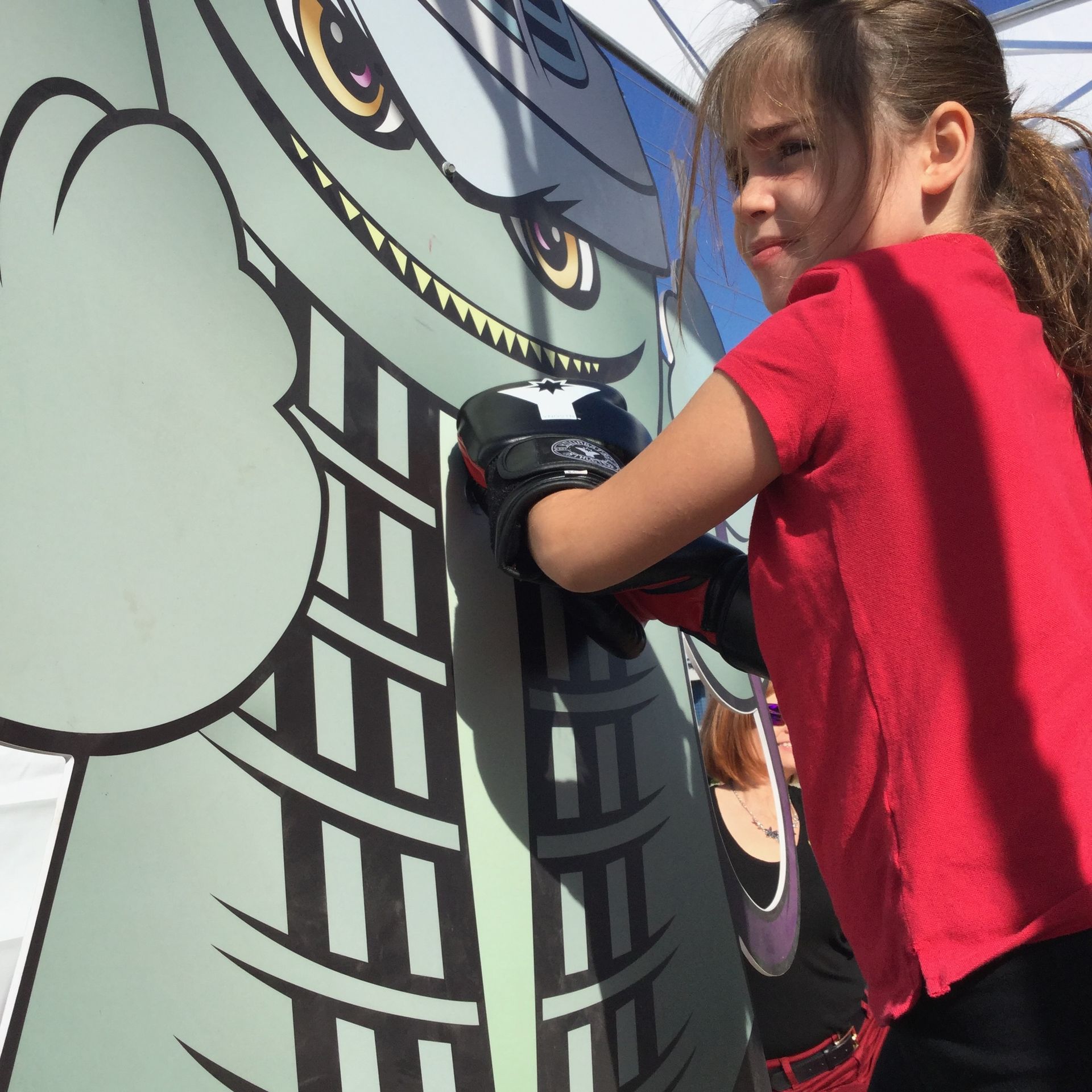 A child wearing boxing gloves punches a large, cartoonish green character cutout standing outdoors.