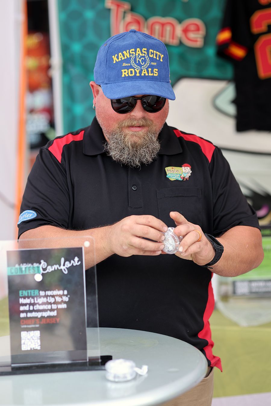 A bearded person in a Kansas City Royals cap and black polo shirt stands at a table, holding a small item near a sign.