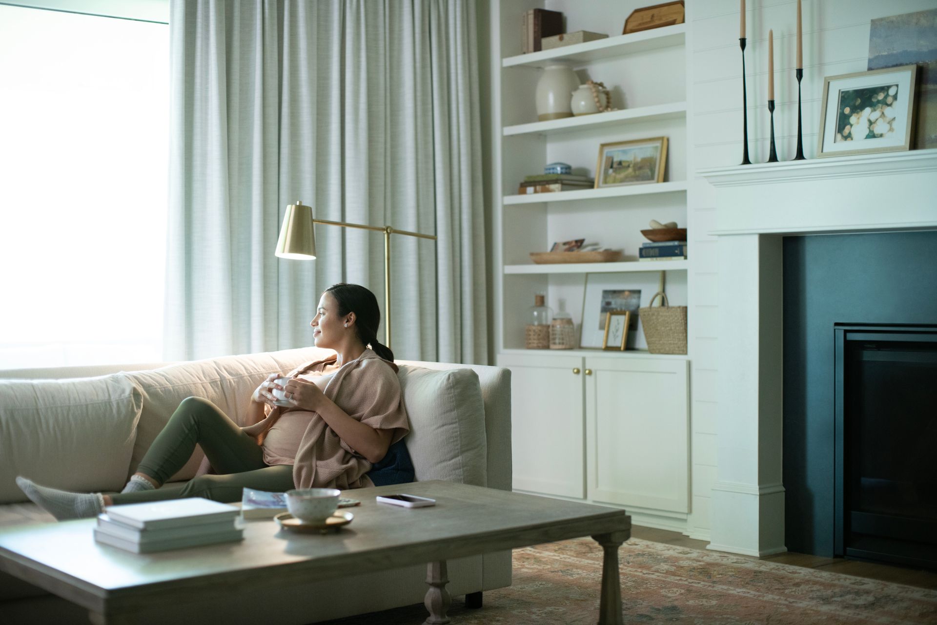 A person rests on a sofa, looking out a window in a sunlit living room with a bookshelf and fireplace.
