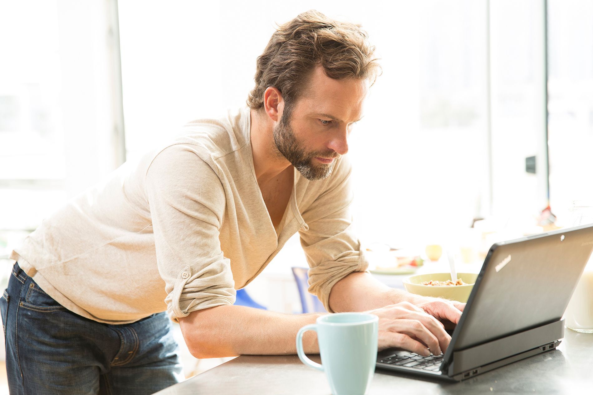 A person in a beige shirt leans over a counter, typing on a laptop next to a light blue mug in a bright, sunlit room.