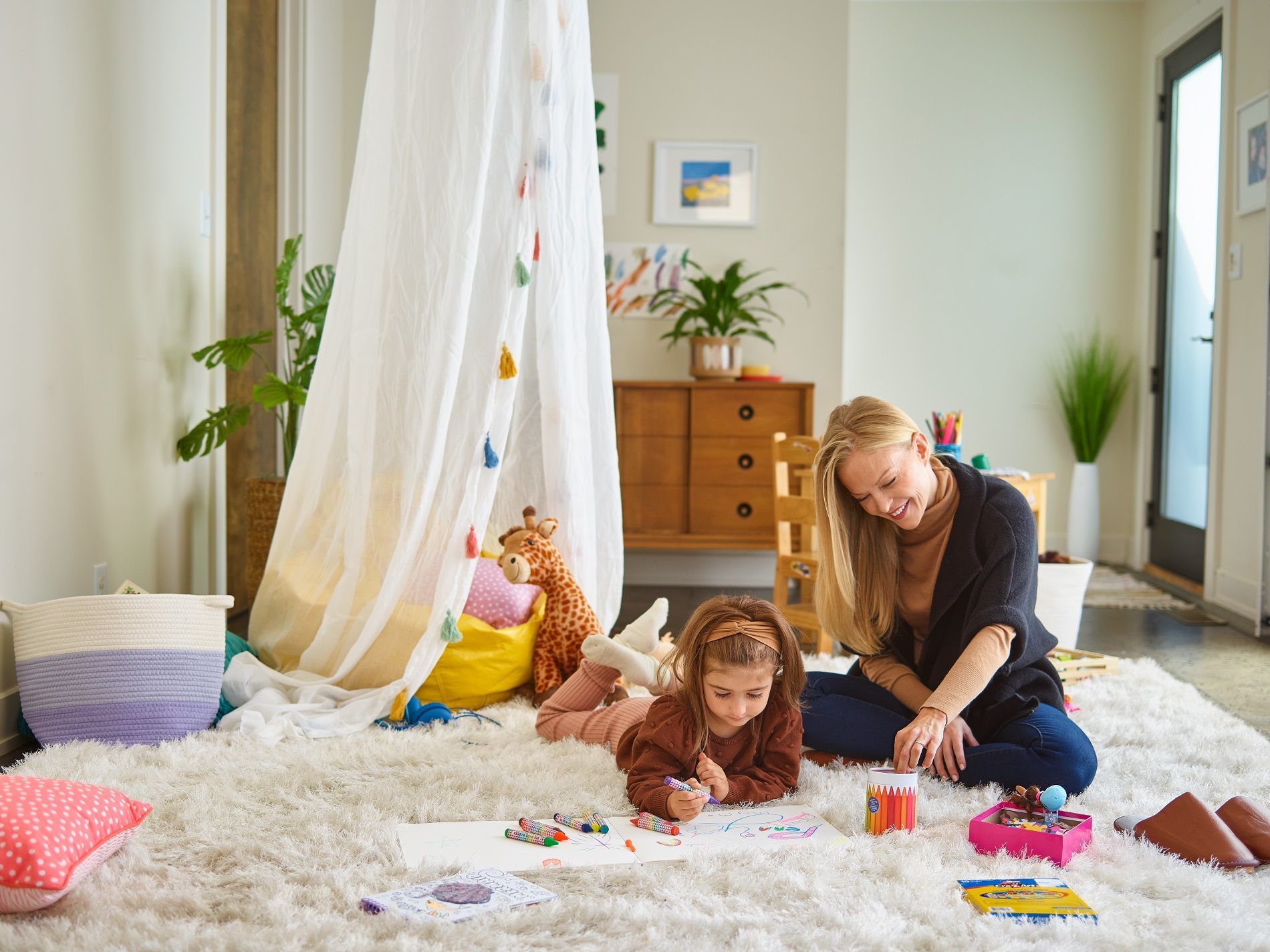 A woman and a child sit on a white rug, coloring together in a cozy room featuring a teepee and wooden furniture.