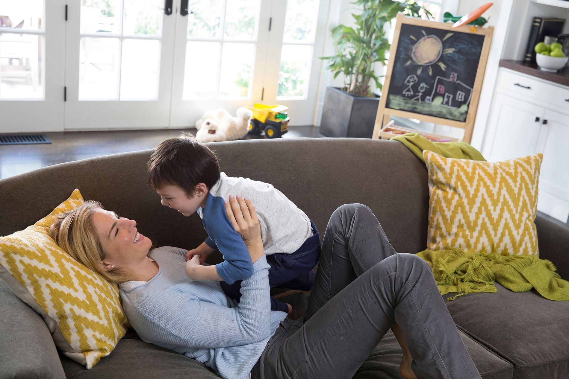 A person lies on a sofa, smiling while interacting with a child sitting on their chest in a bright, modern living room.
