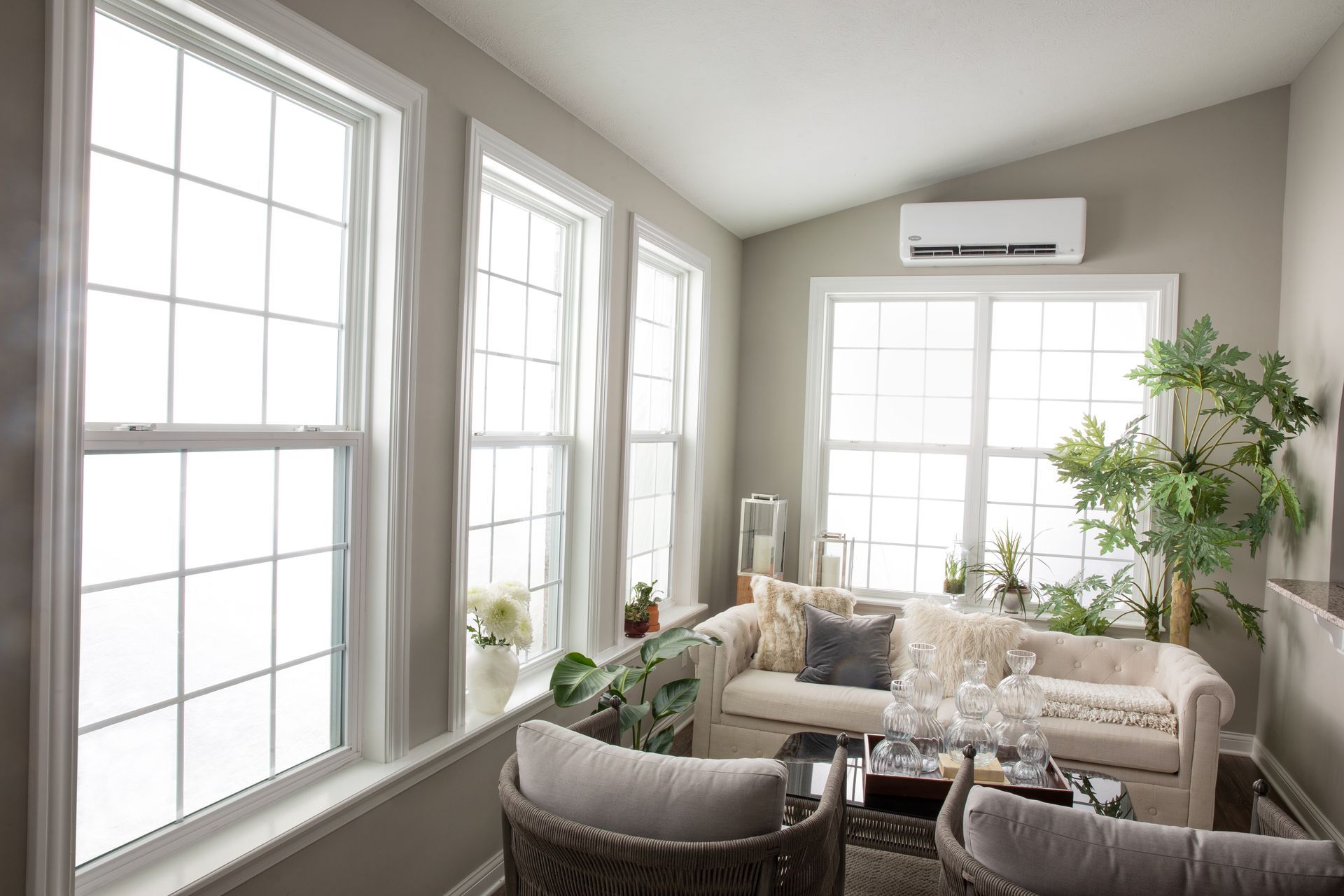 A bright sunroom with light gray walls, large windows, a beige sofa, wicker chairs, and a wall-mounted AC unit.