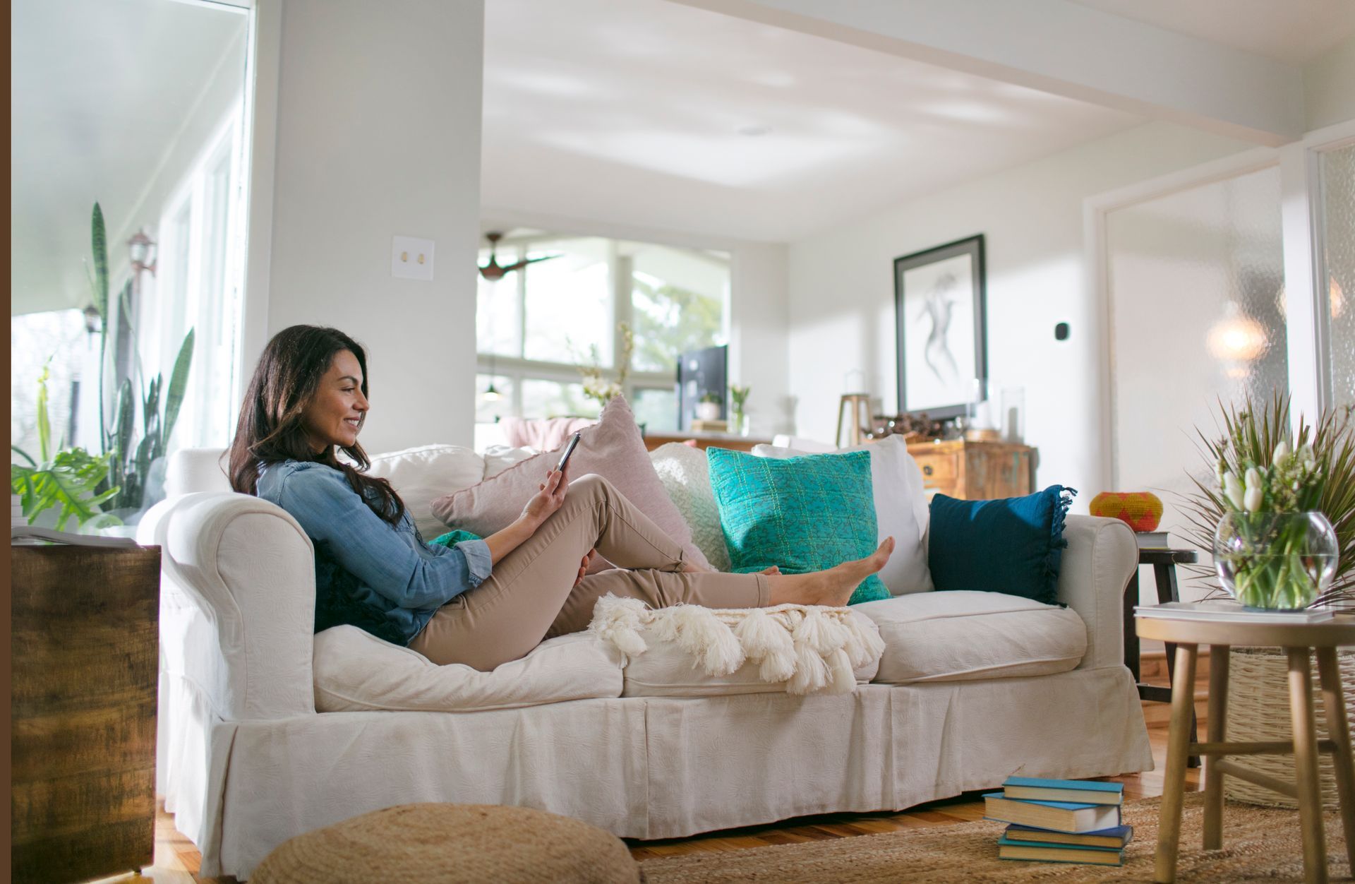 A person relaxes on a white couch in a brightly lit living room, looking at their phone while resting their feet up.
