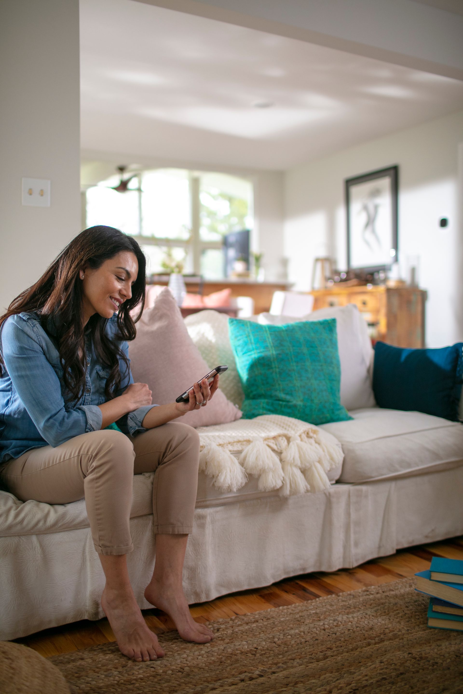 A person sitting barefoot on a white sofa in a living room, smiling while looking at a smartphone held in their hands.