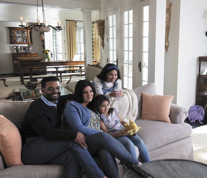 A family sits together on a couch in a bright living room, smiling as they watch television.