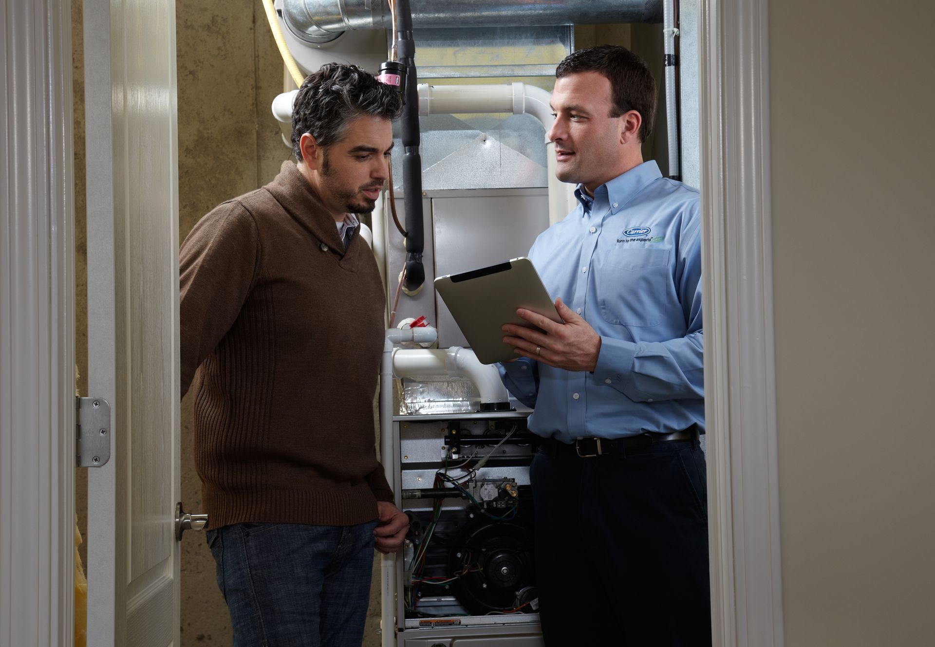 A service technician holding a tablet discusses furnace maintenance with a homeowner in a utility room.