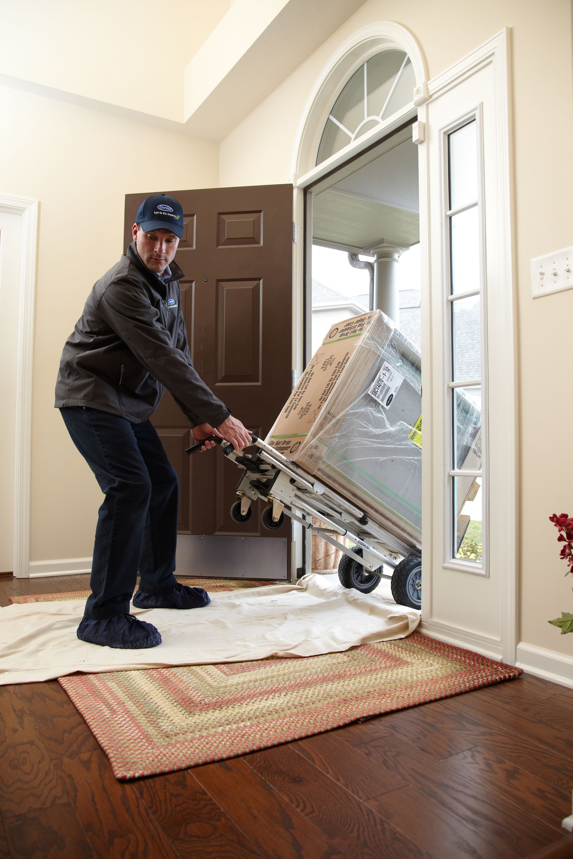 A delivery worker uses a hand truck to move a large, wrapped appliance through an open residential doorway.