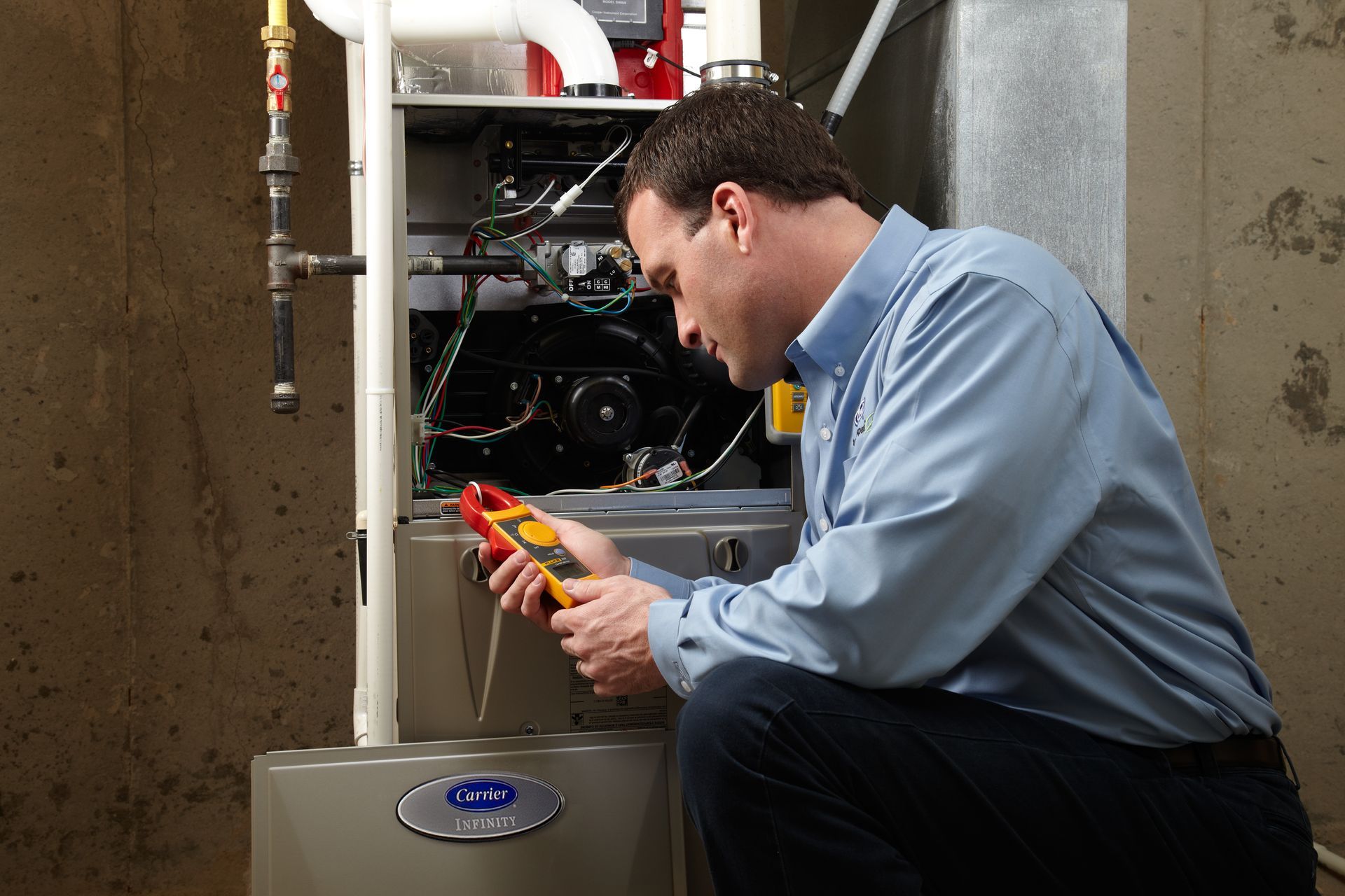 A technician in a blue shirt kneels in a basement, using a multimeter to check the wiring of a heating furnace unit.