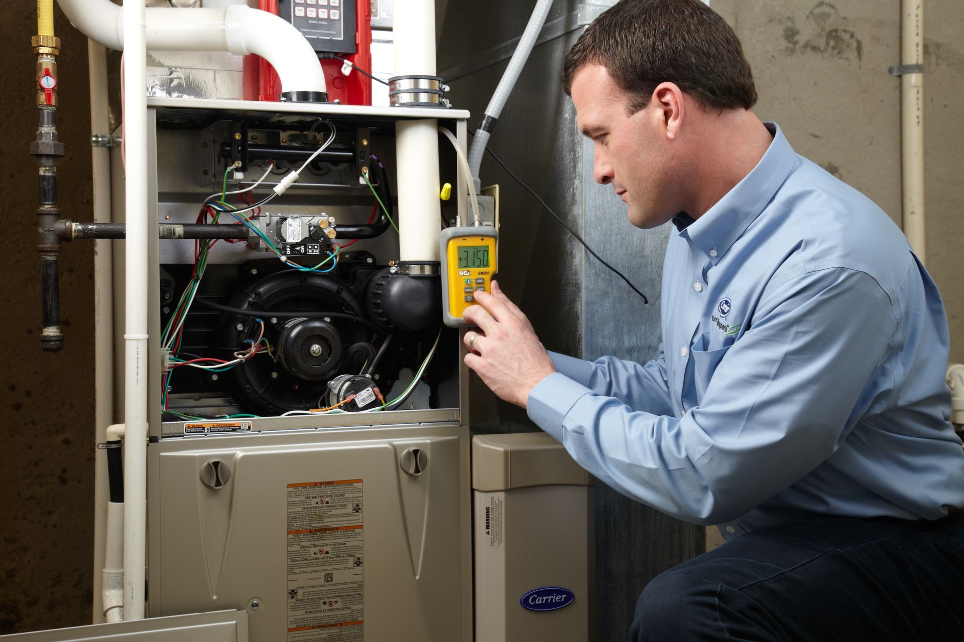 A technician in a blue button-down shirt uses a digital diagnostic tool to inspect an open home furnace unit.