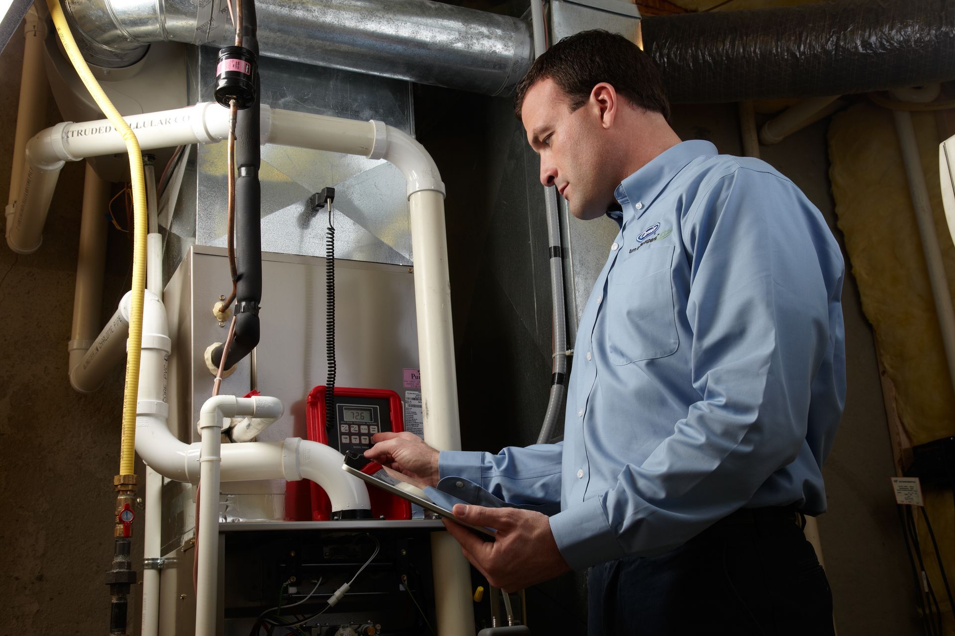 A person in a blue button-down shirt inspects a furnace while holding a clipboard in a utility room.