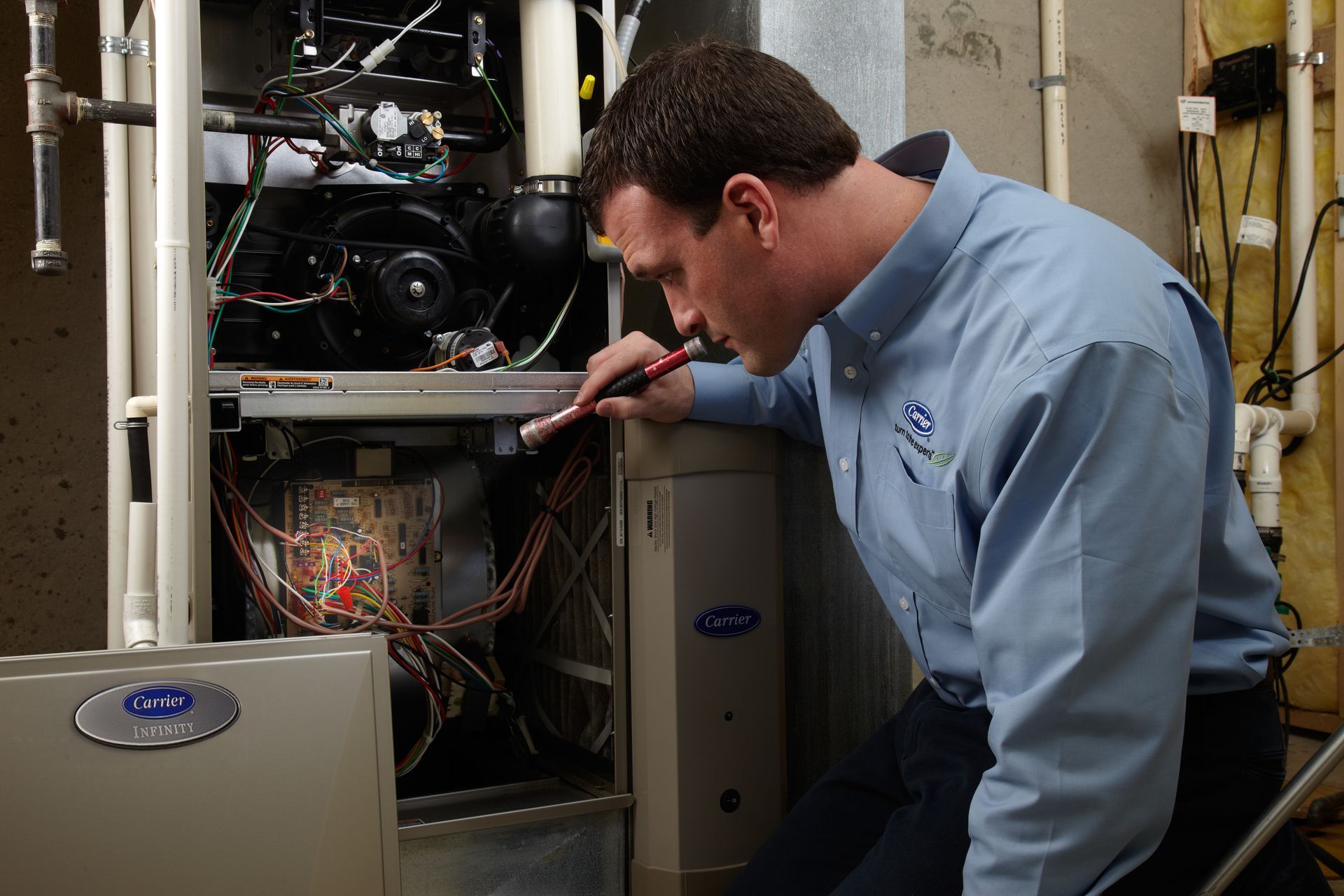 A technician in a light blue shirt uses a flashlight to inspect the interior of an open HVAC furnace unit.