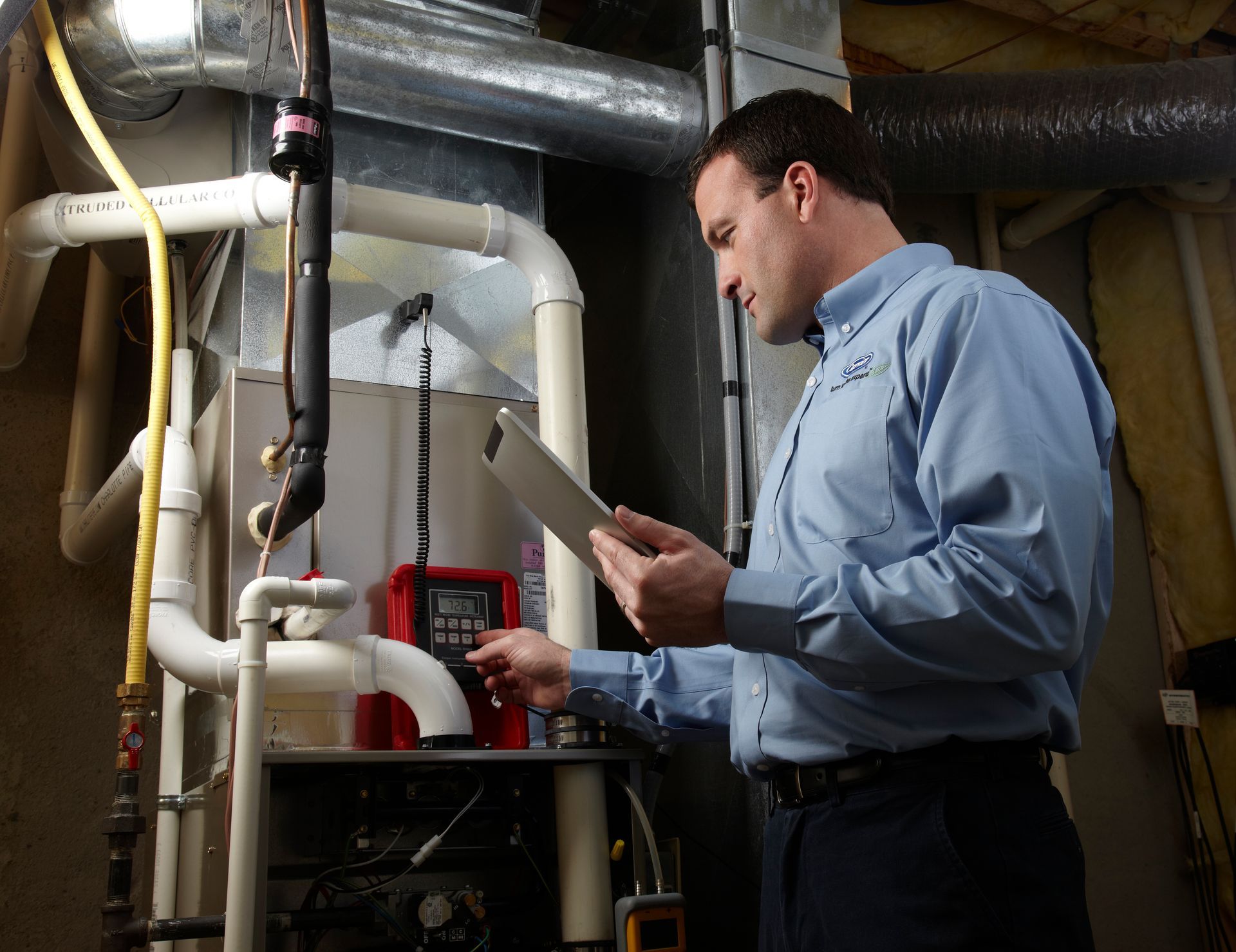 A technician in a light blue shirt inspecting a furnace while holding a tablet in a basement.