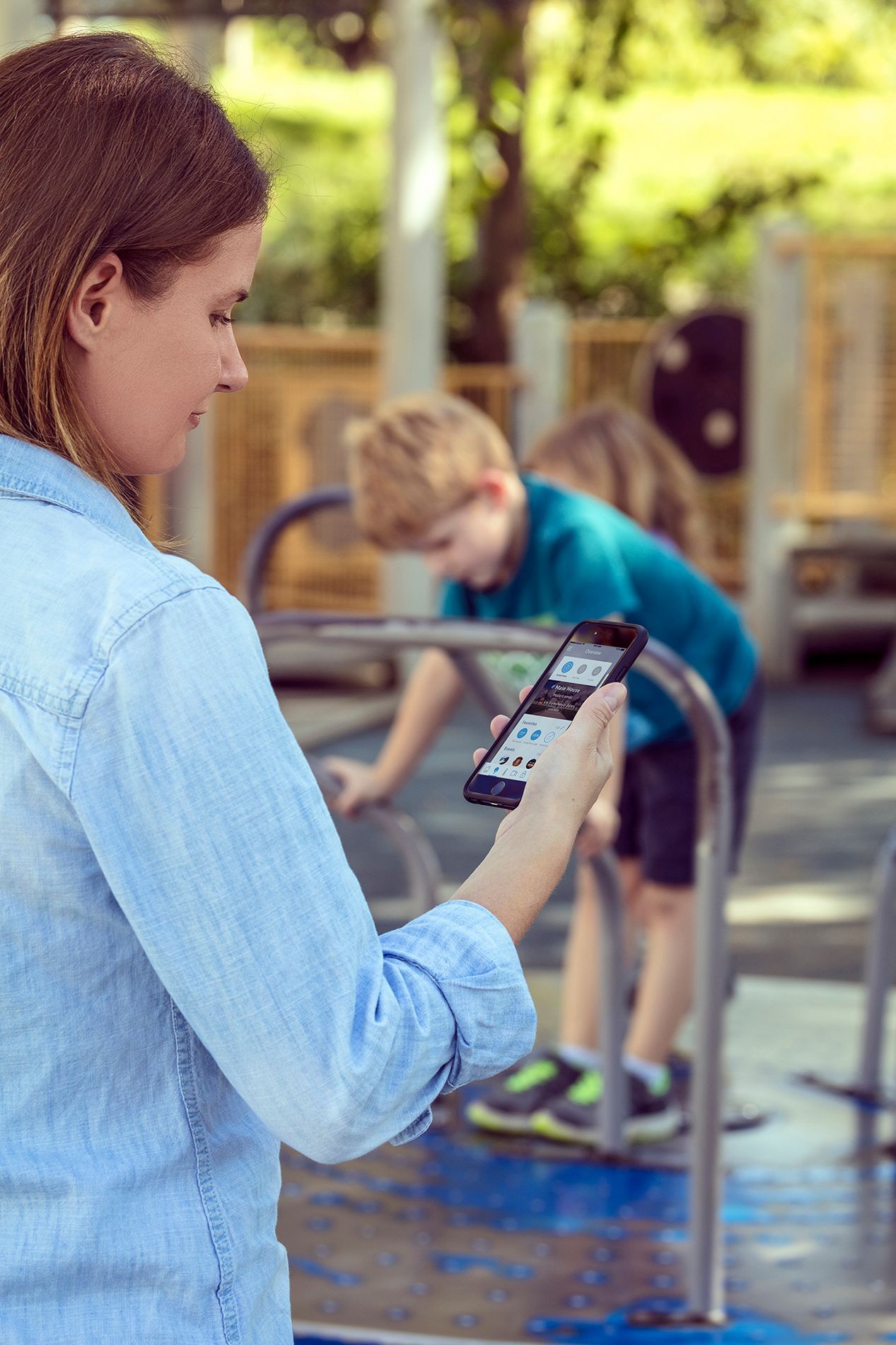 A person in a light blue denim shirt holds a smartphone while observing someone playing on a playground merry-go-round.