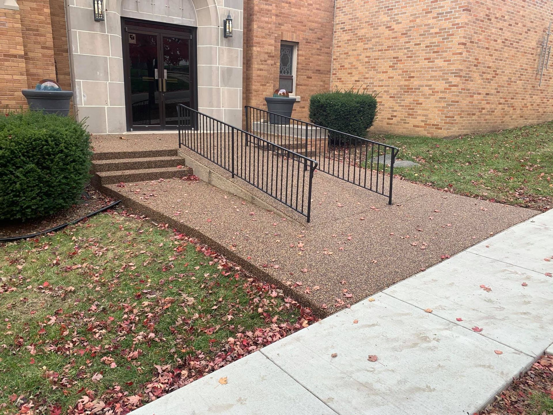 A ramp with black metal handrails leads to a brick building entrance next to a concrete sidewalk and green bushes