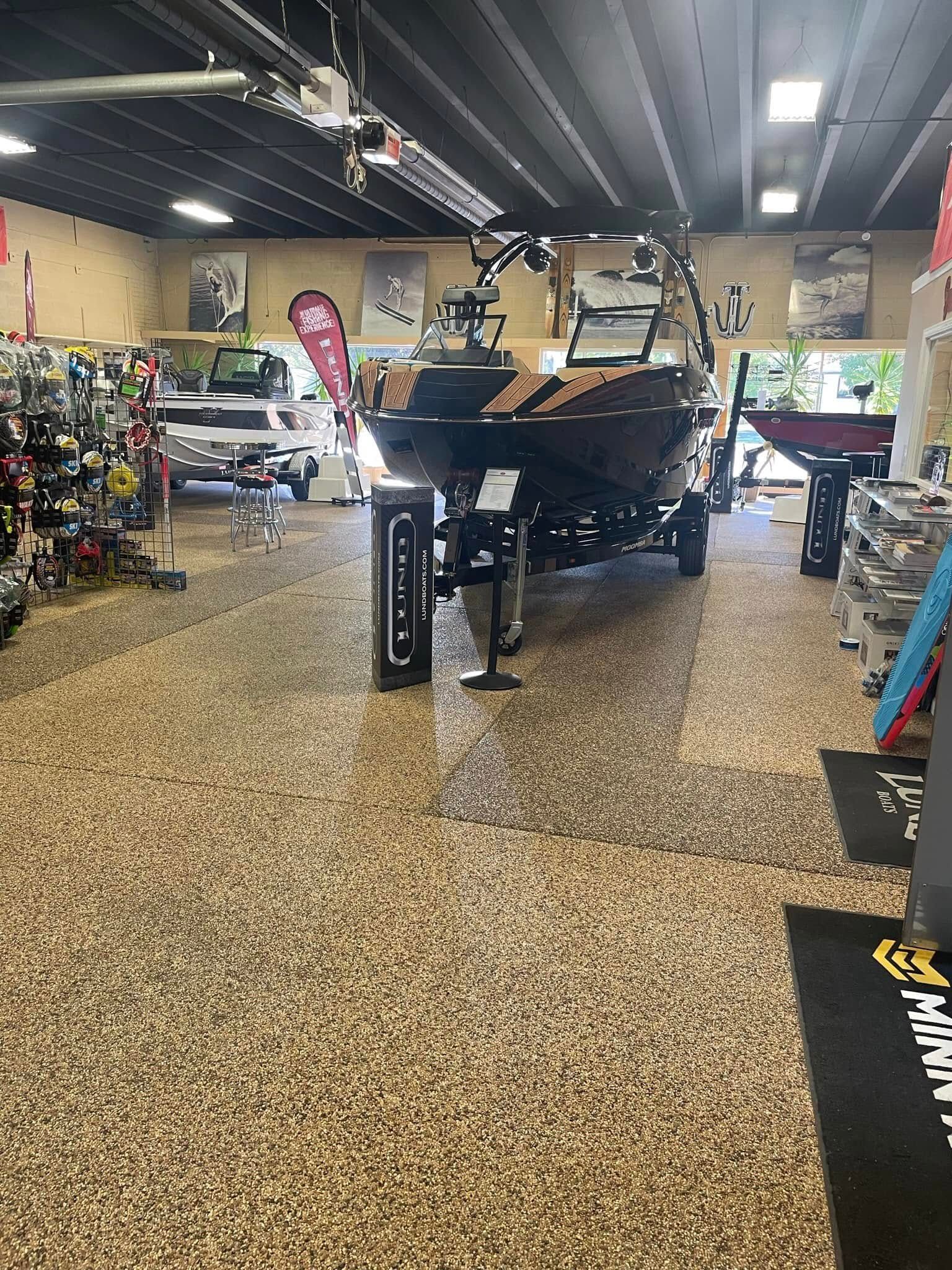 A dark motorboat on a trailer displayed inside a retail store with speckled flooring and various equipment on shelves