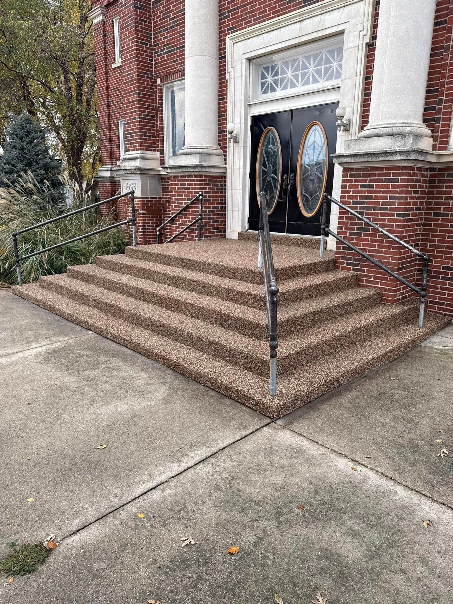 A brick building entrance with five steps covered in a pebble-texture surface, featuring white columns and a black door