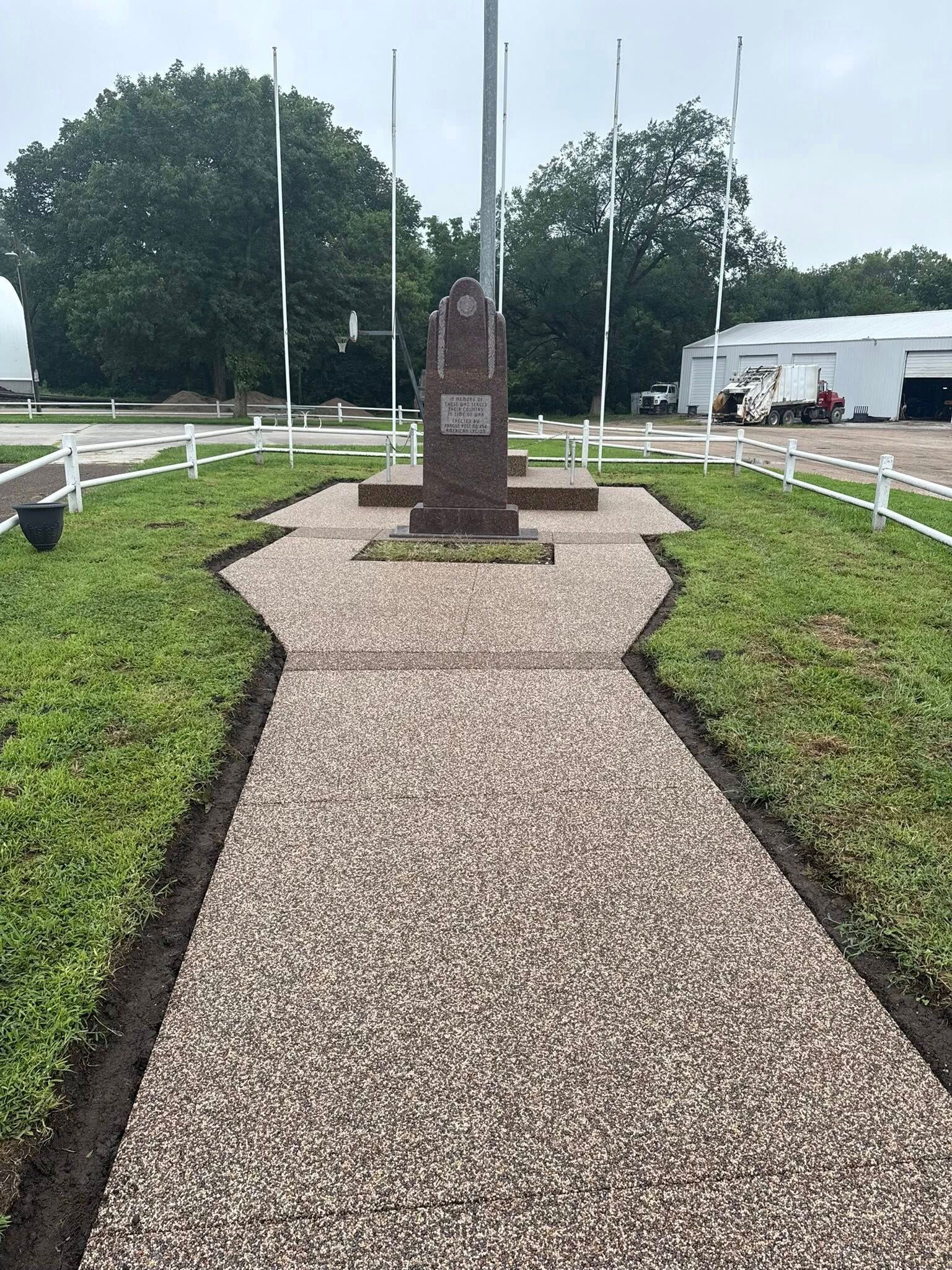 A granite memorial monument stands at the end of a stone path in a grassy area surrounded by white fence rails and poles