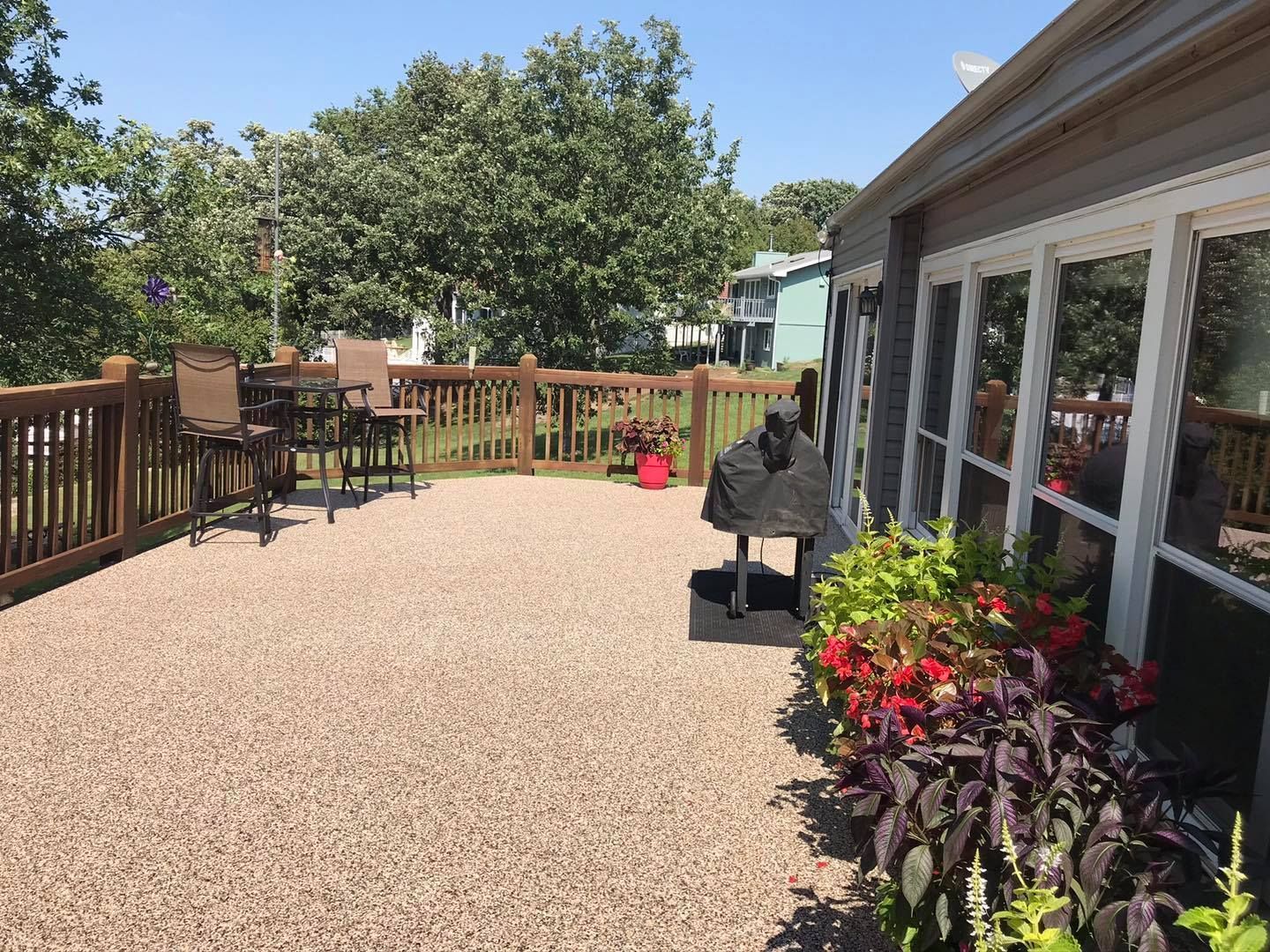 A sunny backyard deck featuring tan gravel flooring, wooden railings, outdoor seating, a grill, and vibrant flower beds